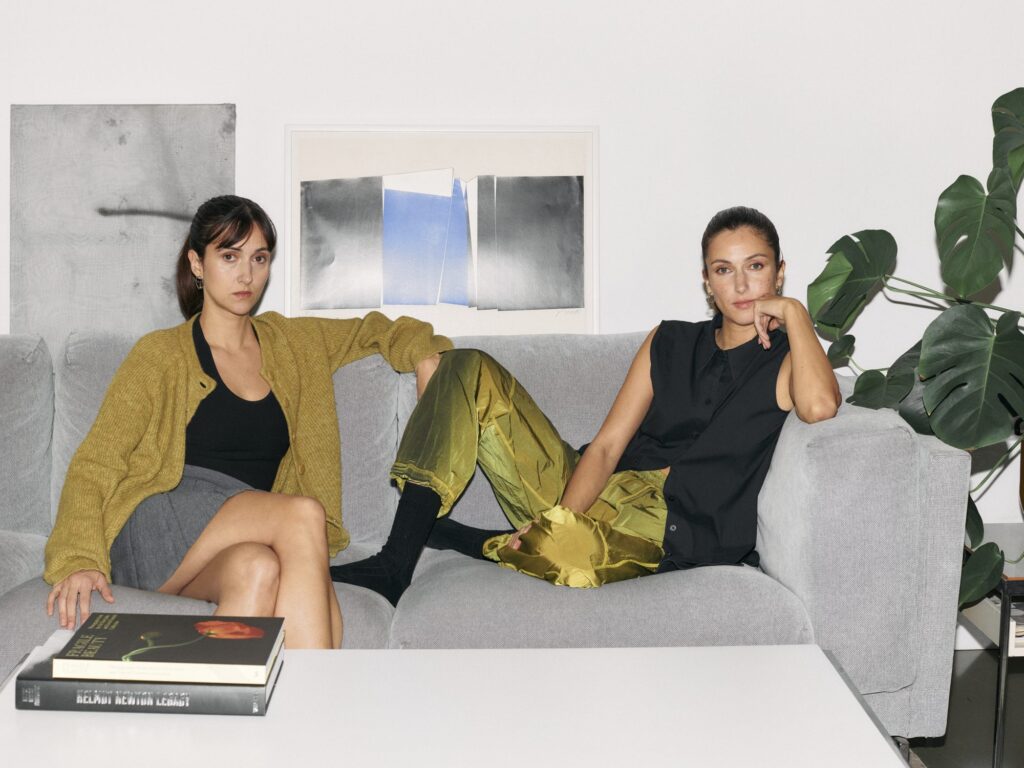 Portrait of Flo and Kim Nüesch seated on a light grey sofa in a contemporary living room, wearing coordinated black and olive green outfits.