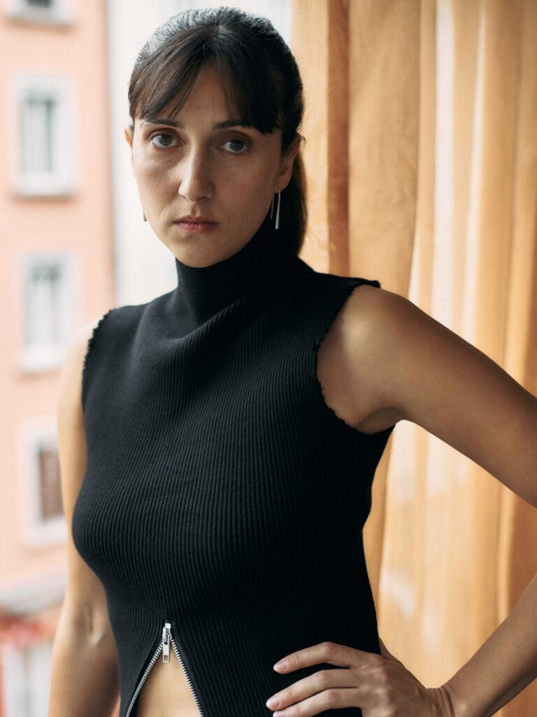 Portrait of Kim Nüesch, wearing a black sleeveless ribbed knit top and silver earrings, standing outdoors against a softly blurred urban background.