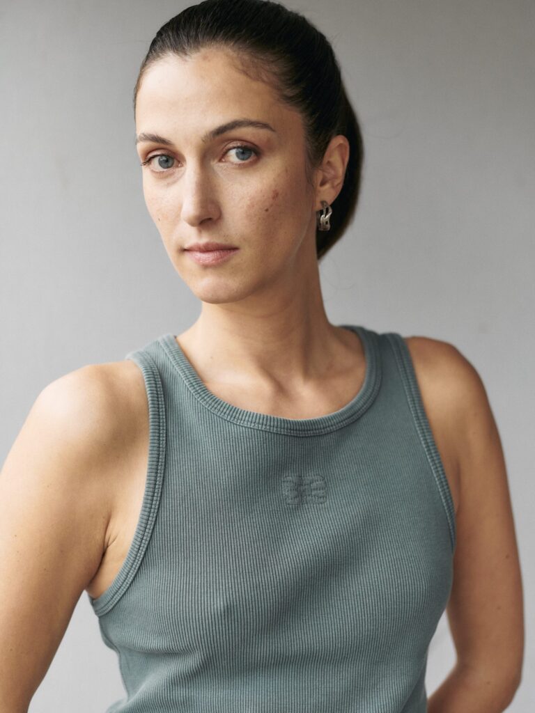 Portrait of Kim Nüesch, wearing a sage green ribbed tank top and silver earrings, looking directly at the camera against a neutral background.