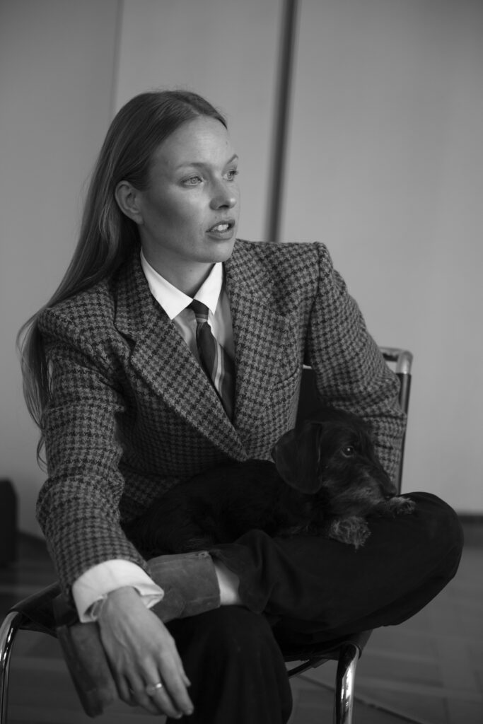 Woman wearing a checked blazer and tie, seated with a dachshund resting on her lap.