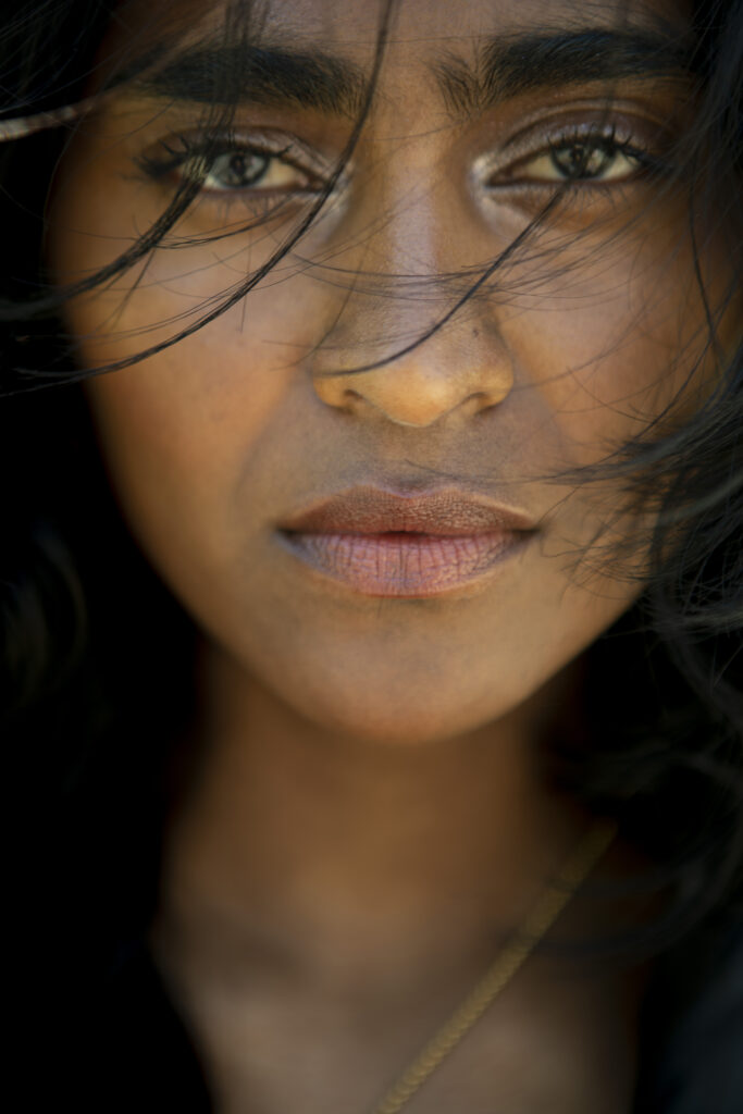 Close-up beauty portrait of model Barathy with wind-swept dark hair framing her face, soft natural light and a direct, calm gaze.