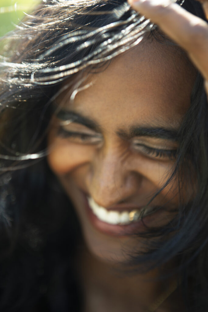 Close-up of a smiling person with long dark hair, gently running a hand through their hair in natural sunlight.