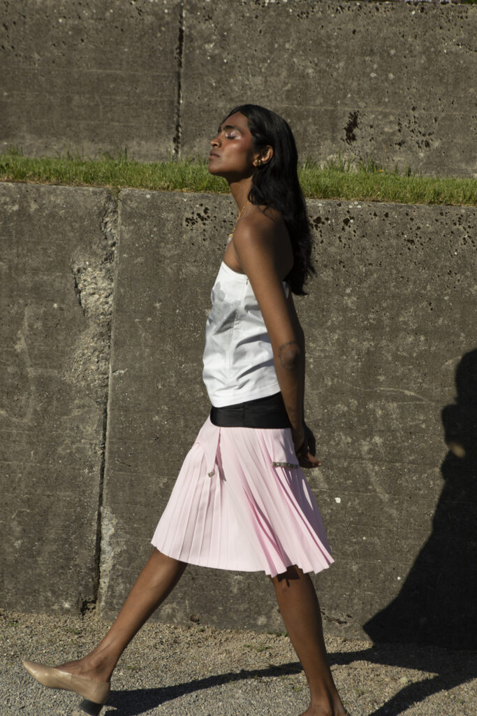 Barathy Akkan walking in profile wearing a white top and a pale pink pleated skirt in bright afternoon sunlight.