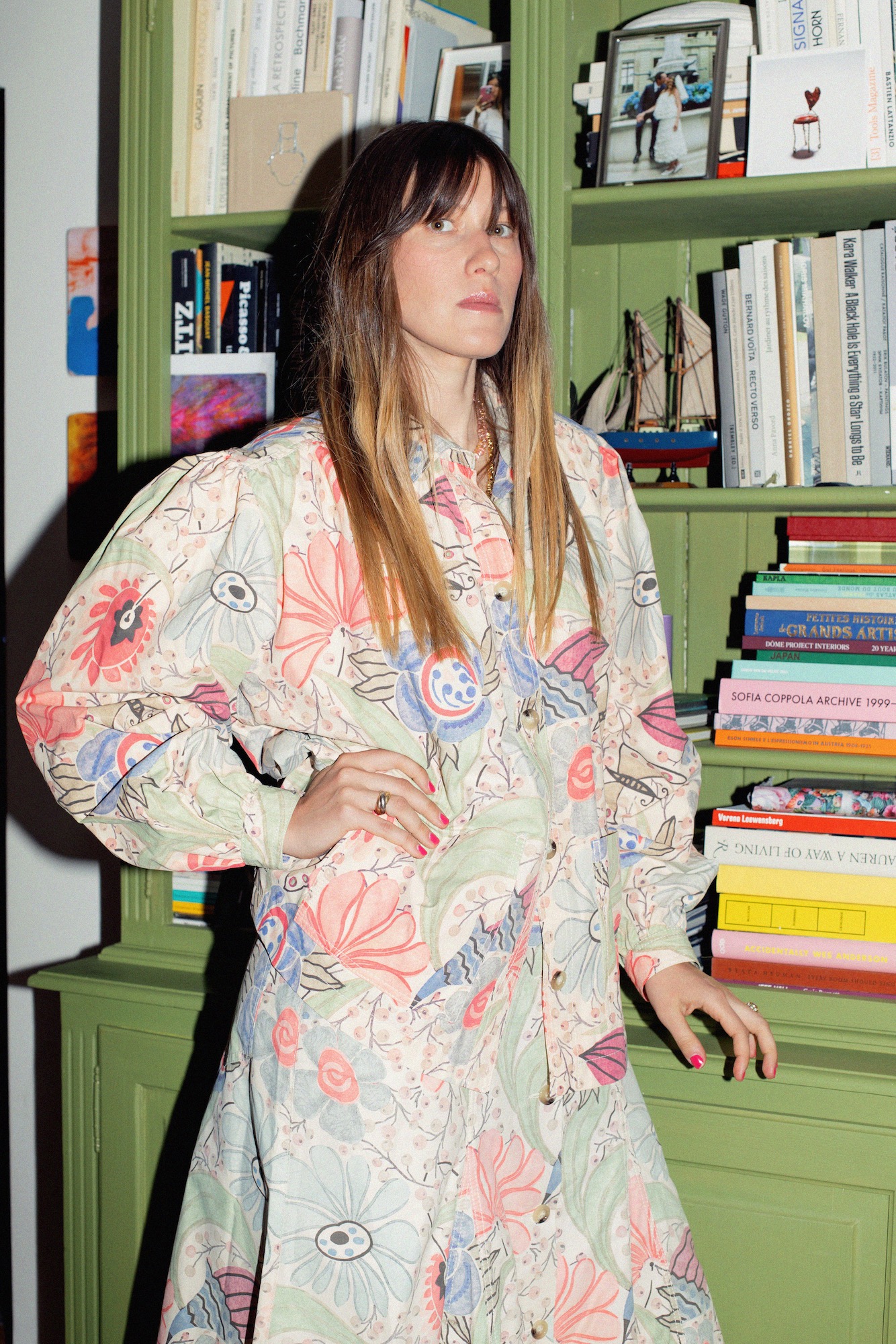 Portrait of Anouck Mutsaerts beside a green bookshelf, wearing a patterned outfit and layered jewellery.