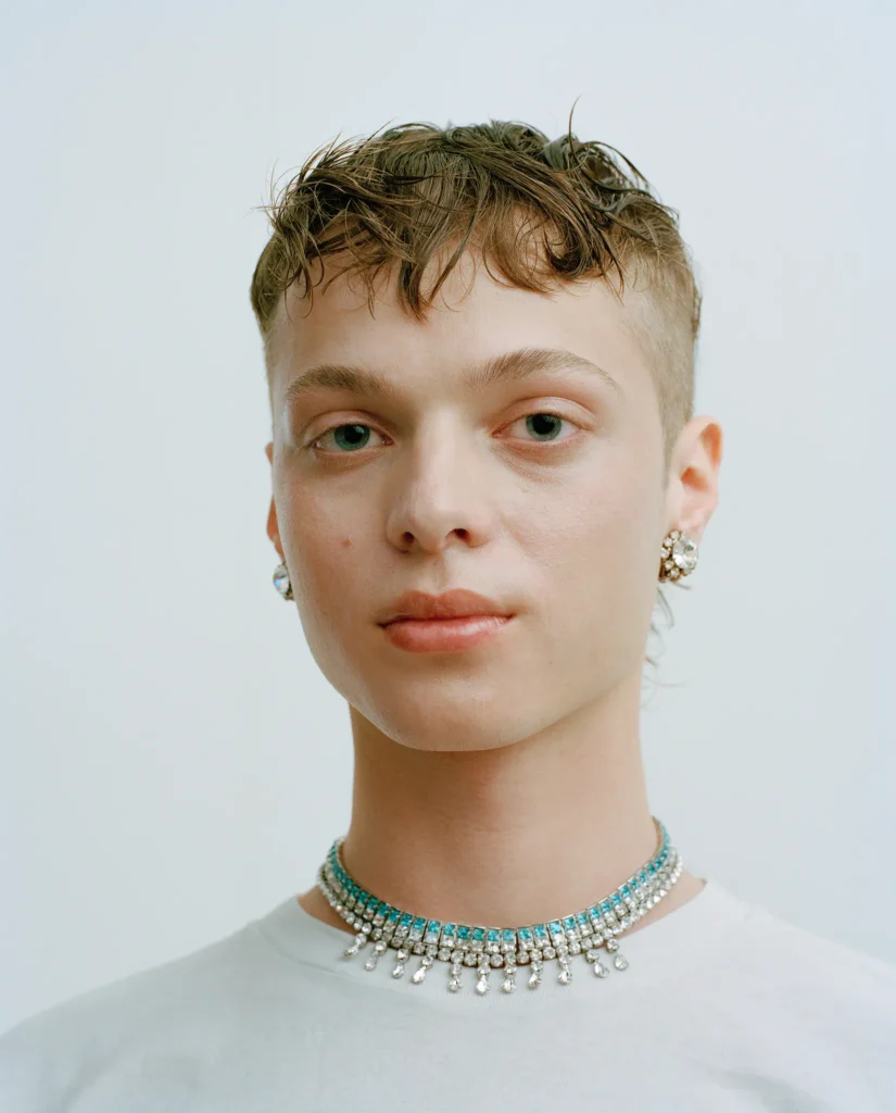 Portrait of Béla with short tousled hair, wearing a white T-shirt and turquoise necklace.