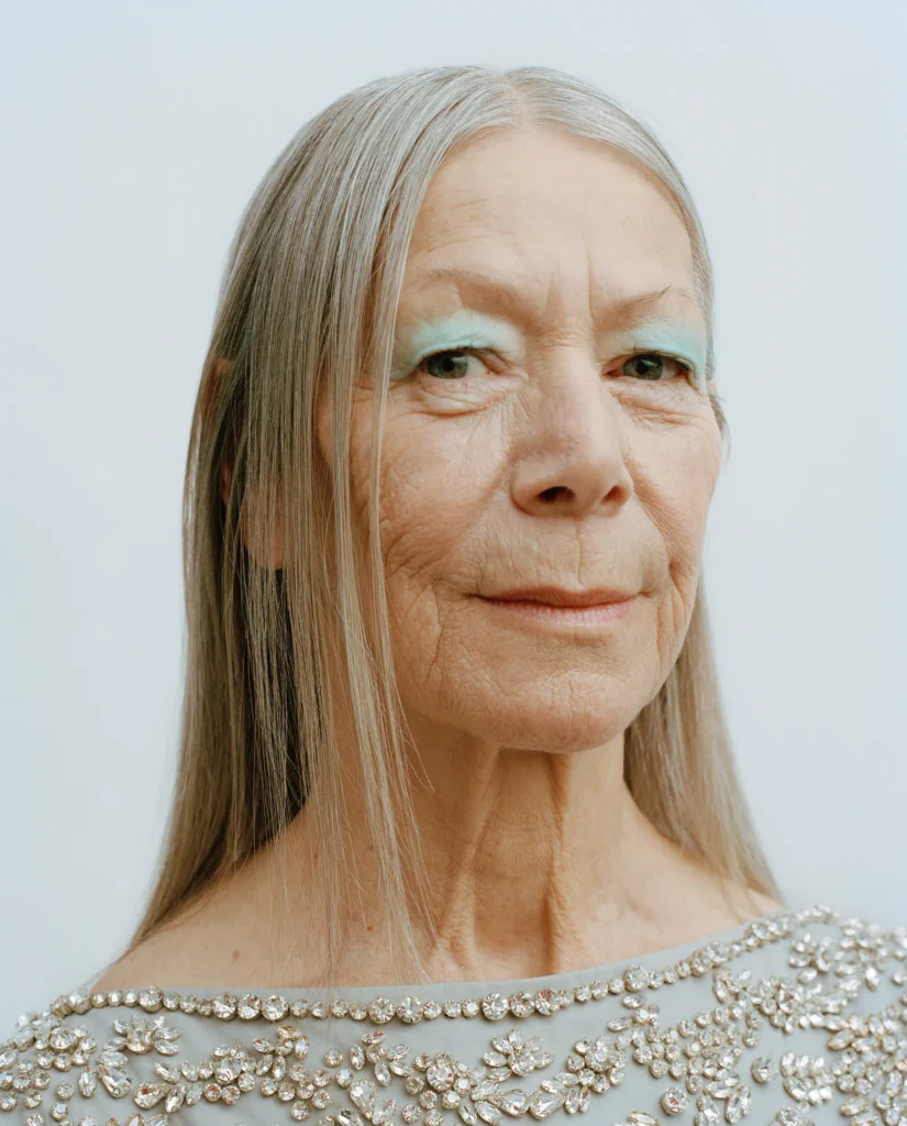 Portrait of Claudia with long straight grey hair, pale green eye makeup and an embellished top.
