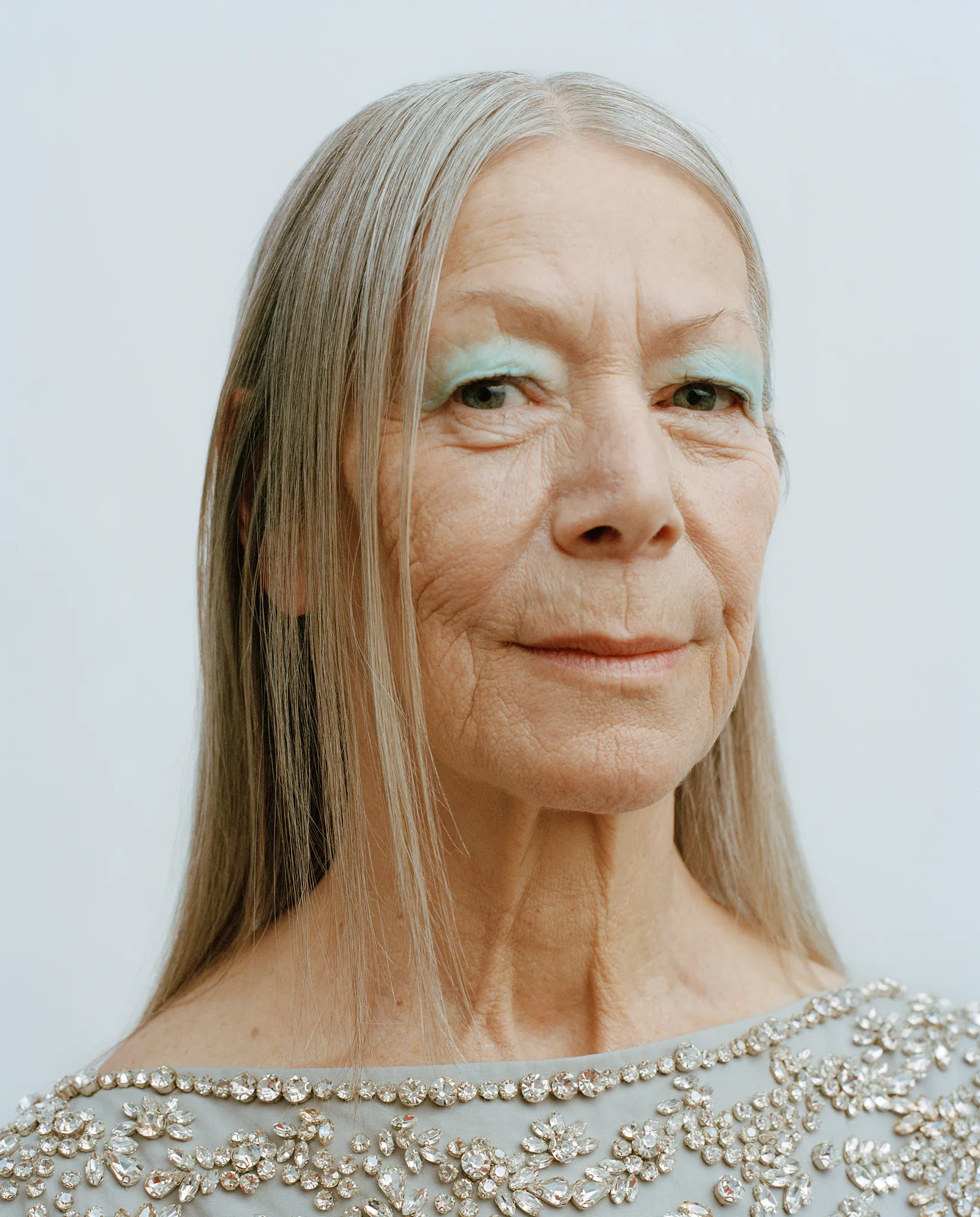 Portrait of Claudia with long straight grey hair, pale green eye makeup and an embellished top.
