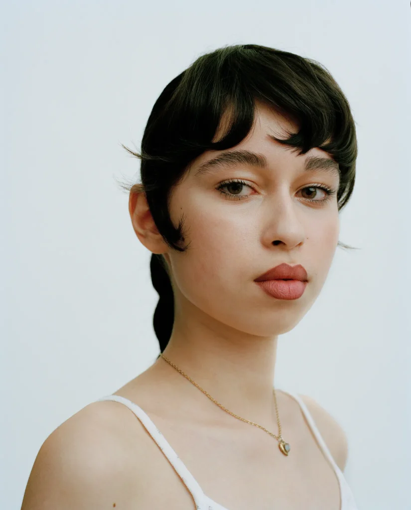 Portrait of Jasmina with short dark hair, wearing a white camisole and delicate necklace against a pale background.