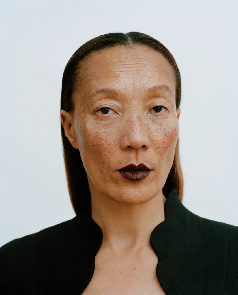 Portrait of Kojolee with slicked-back hair, freckles and dark lipstick, wearing a black jacket.