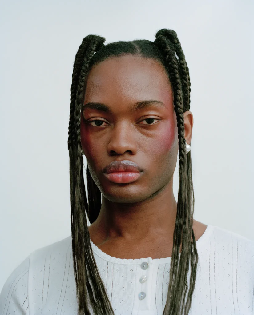 Portrait of Pharrell with long braided hair, wearing a white top against a pale background.