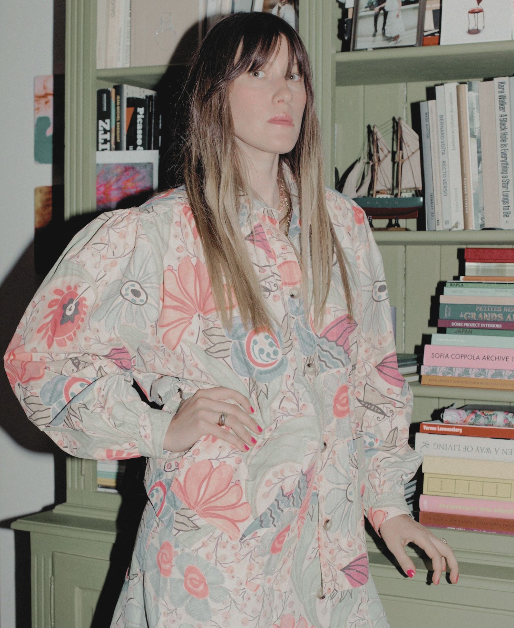 Anouck Mutsaerts dans une tenue fleurie, debout à côté d’une bibliothèque verte garnie de livres, d’œuvres d’art et d’objets collectionnés.
