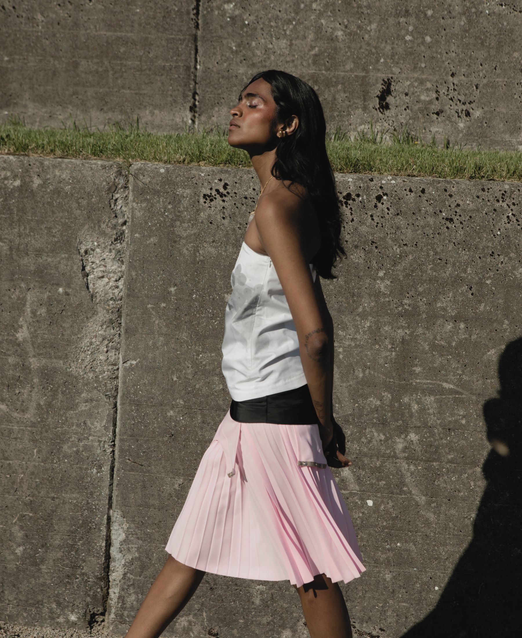 Barathy Akkan in a white top and pleated pink skirt, standing in profile against a raw concrete wall.