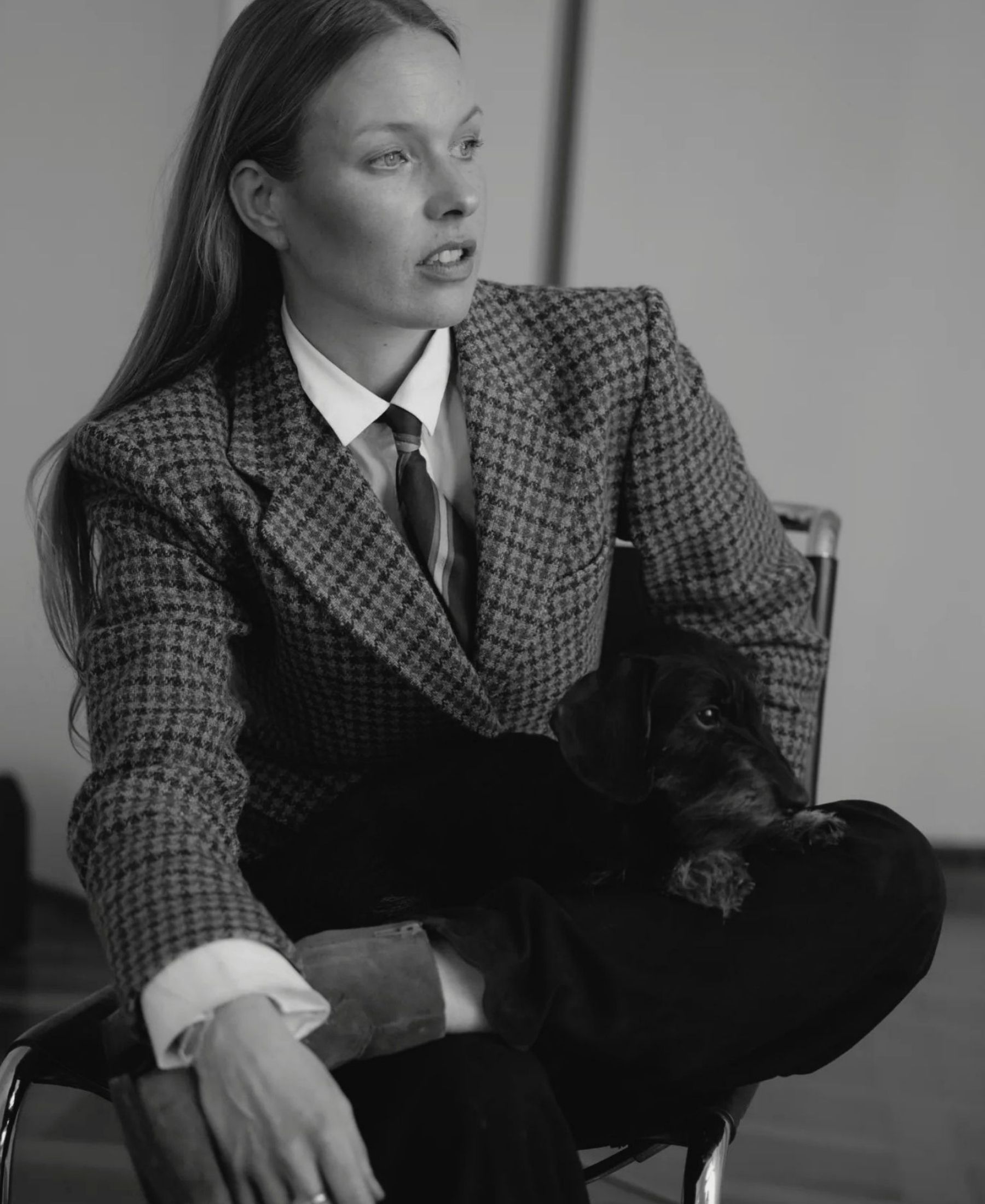 Raphaela Pichler seated in a houndstooth blazer and tie, with a small black dachshund resting across her lap.