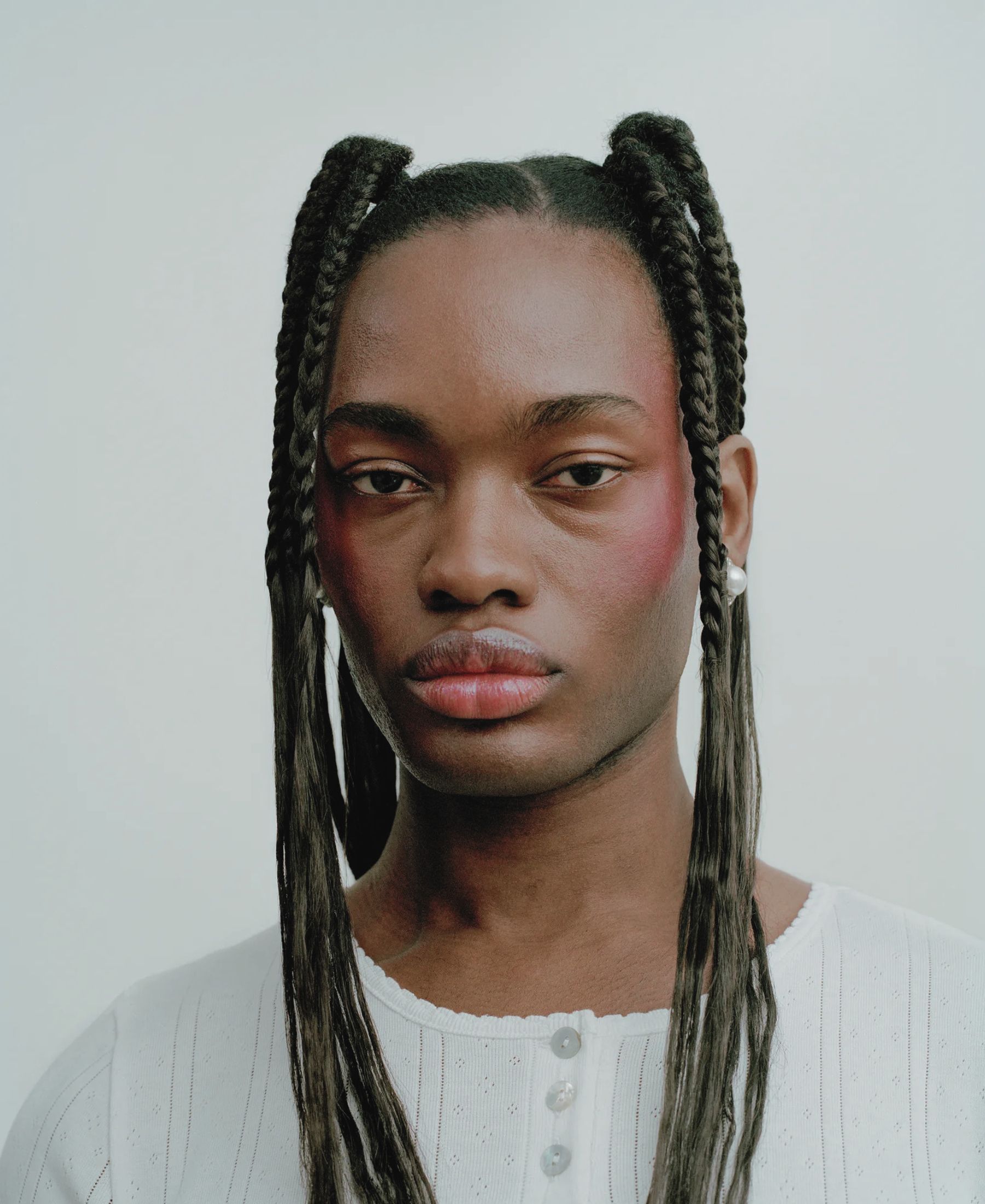 Pharell in close-up, with long sculpted braids, softly flushed cheeks and pearl earrings.