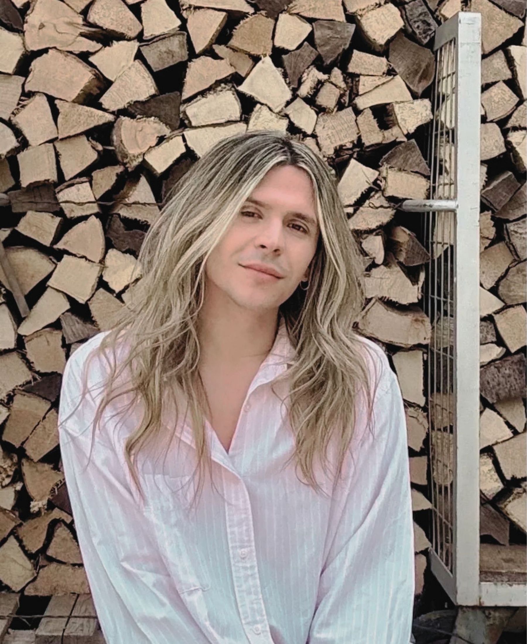 Philipp Rossmann with long light hair and a pale striped shirt, photographed in front of a stack of chopped wood.