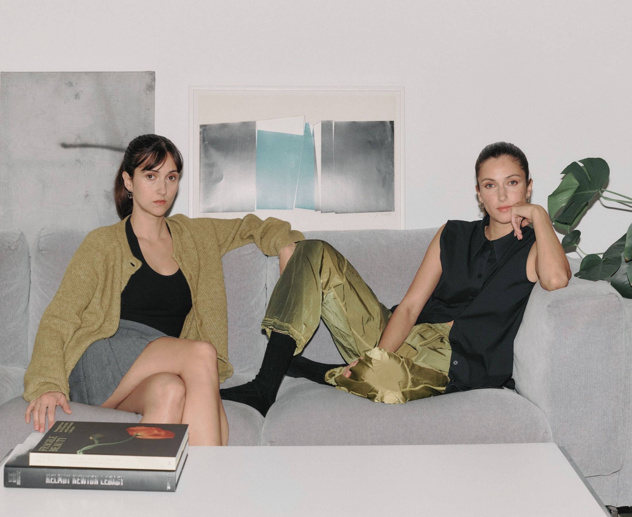 Florine and Kim Nüesch seated at opposite ends of a grey sofa, surrounded by art, books and a large green plant.