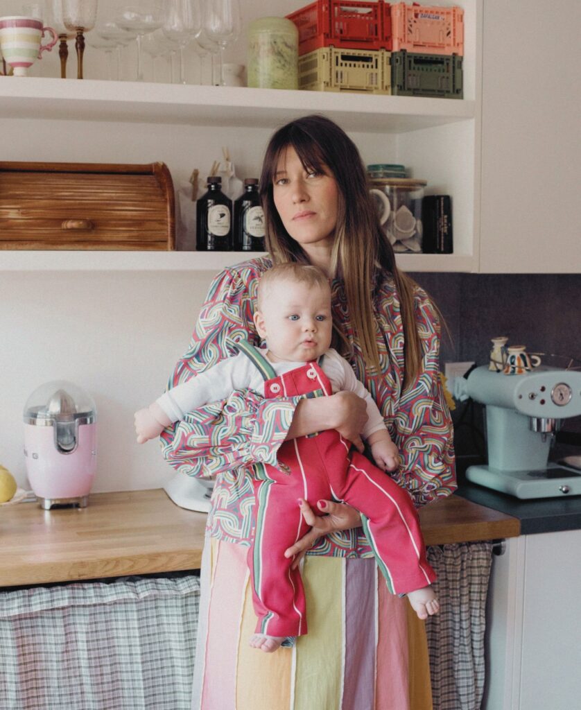 Anouck Mutsaerts hält ihr Baby in der Küche im Arm, umgeben von farbenfrohen Alltagsgegenständen.