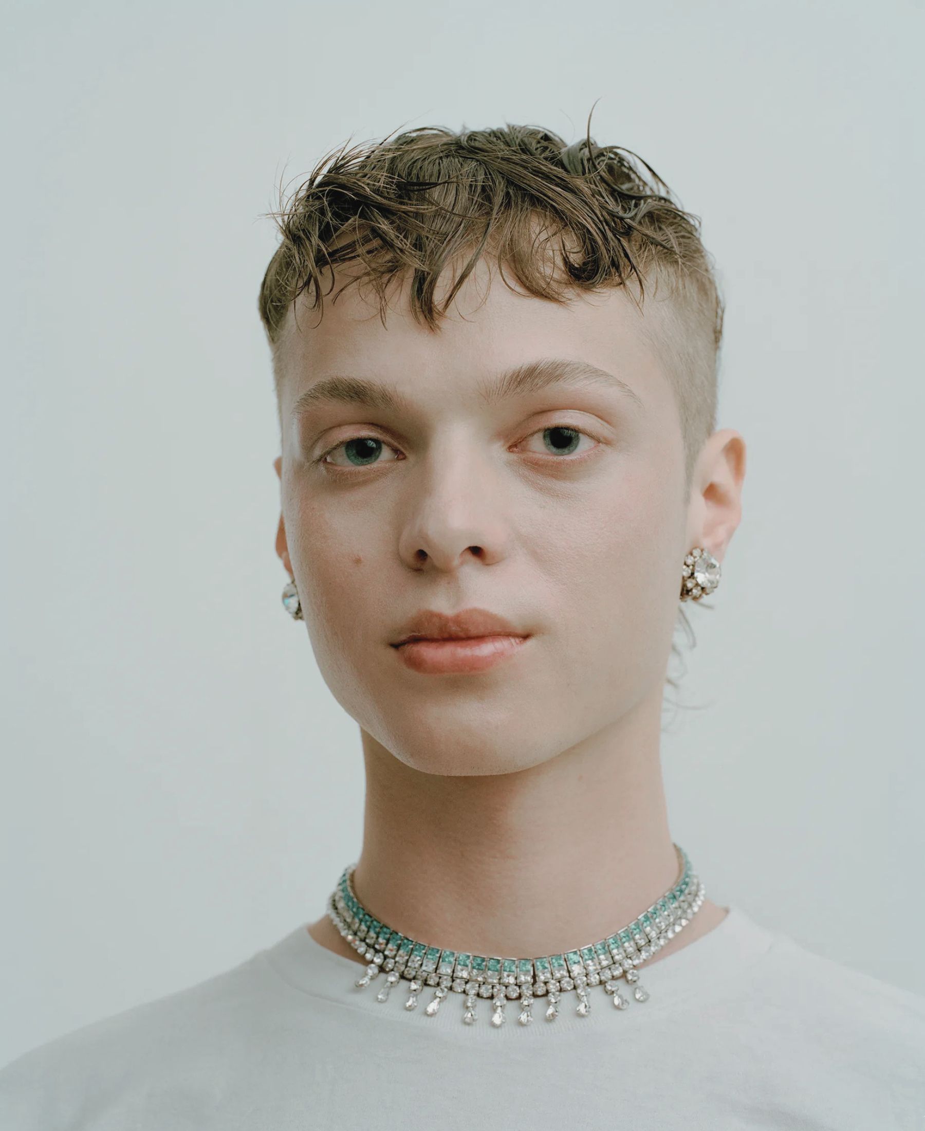 Béla in close-up, with cropped wet-look hair, crystal earrings and layered pale blue and silver necklaces.