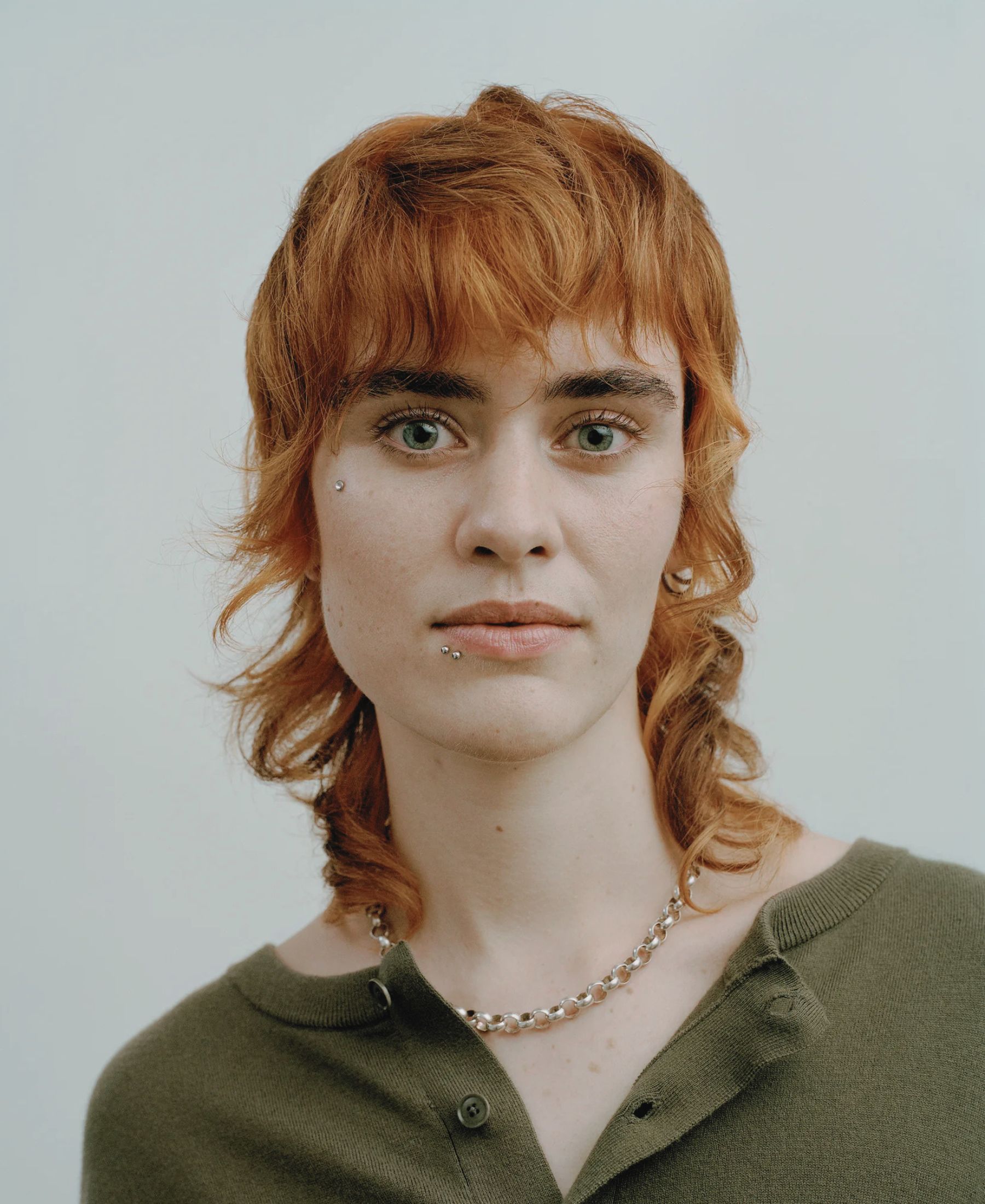 Tosca in close-up, with copper-red hair, subtle facial piercings and a dark olive cashmere top.