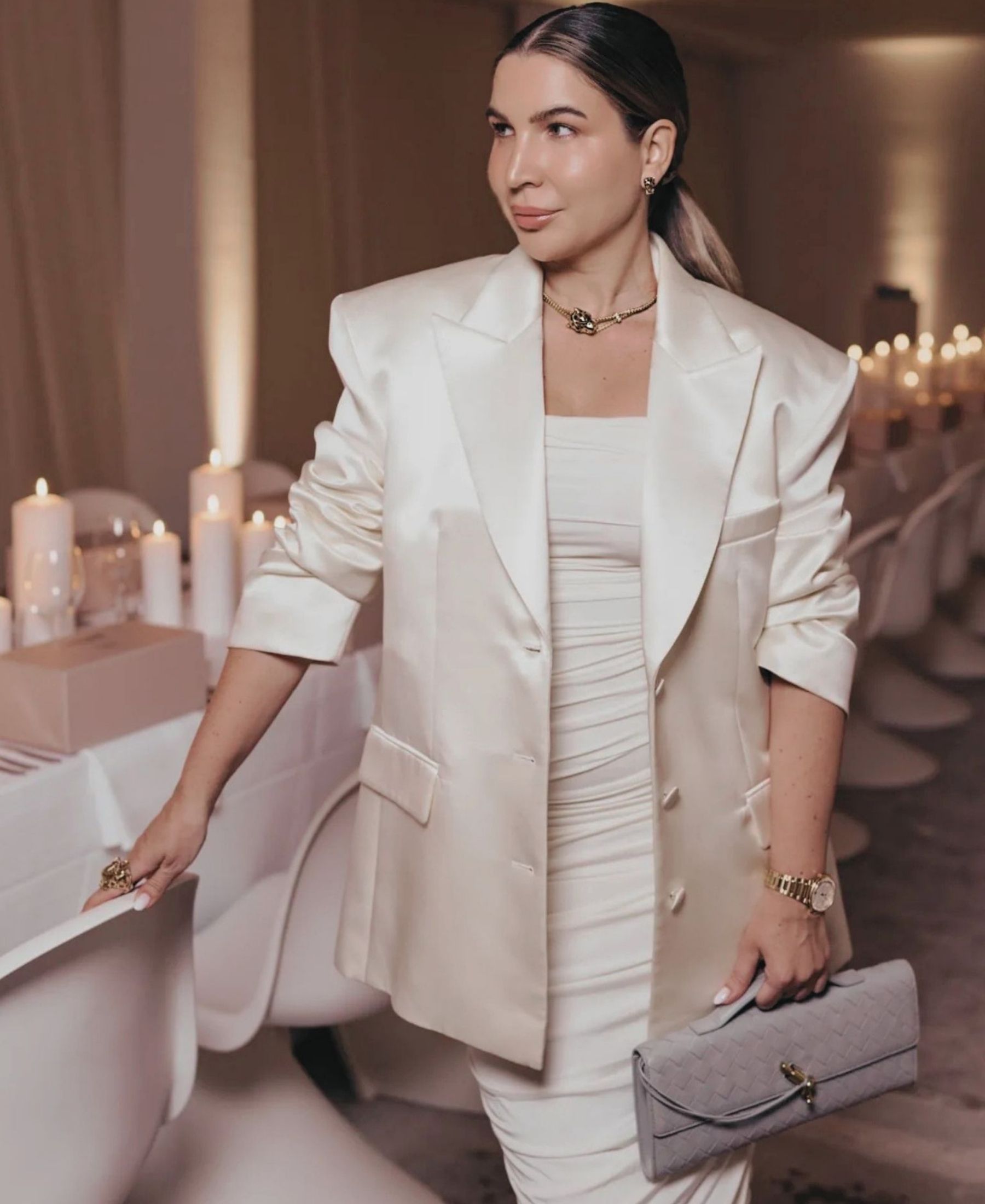 Leyla Hajrovic in a white tailored look, holding a pale woven clutch in a softly lit interior.