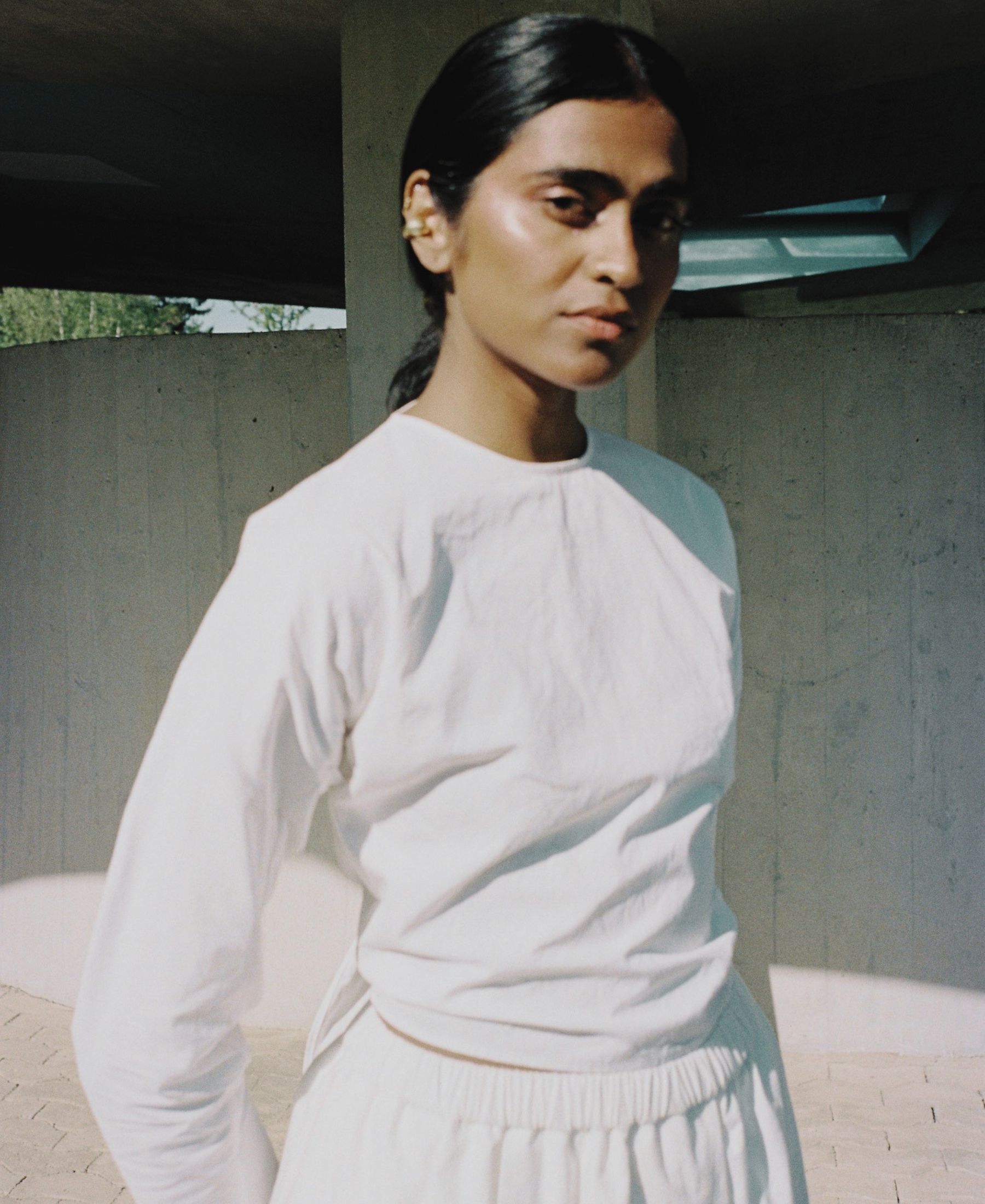 Barathy Akkan in a sculptural white look, standing beneath a concrete structure in sharp afternoon light.
