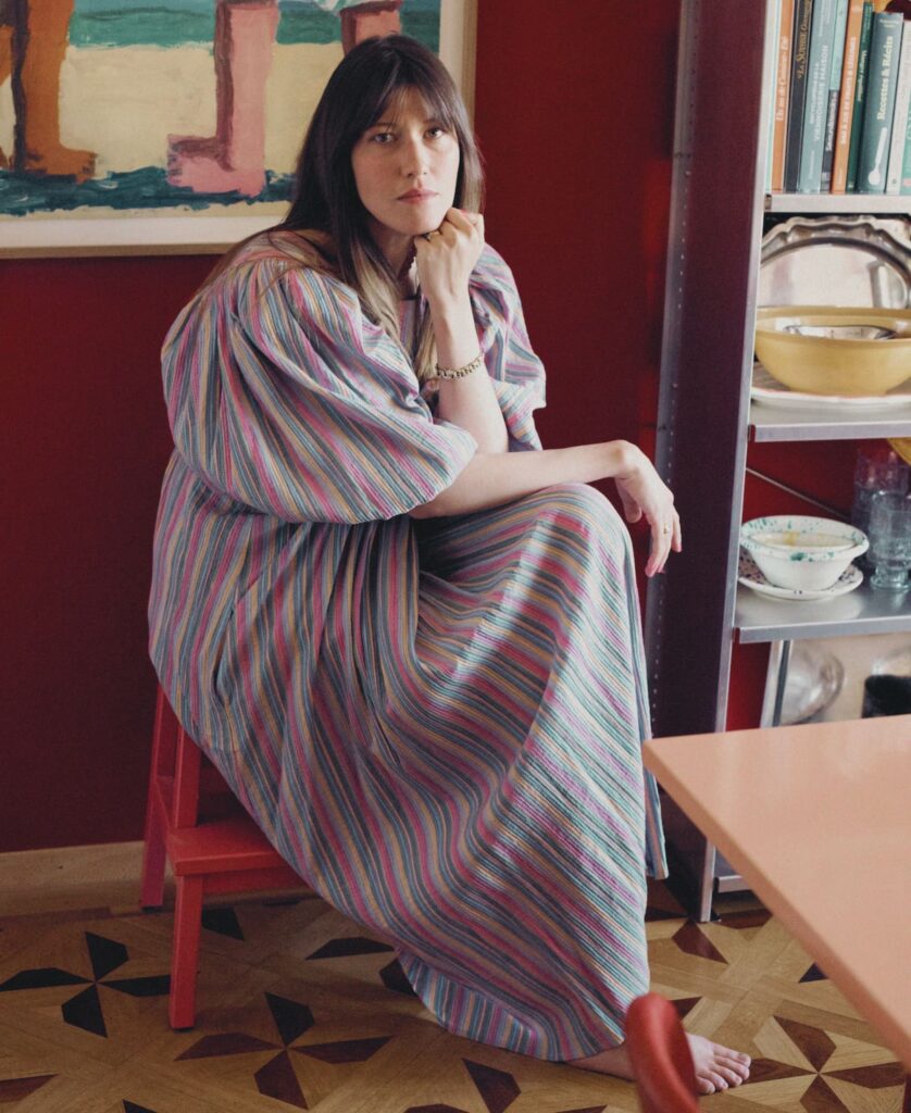 Anouck Mutsaerts sitzt in einem gestreiften Kleid vor tiefroten Wänden, umgeben von Büchern, Keramik und Kunst