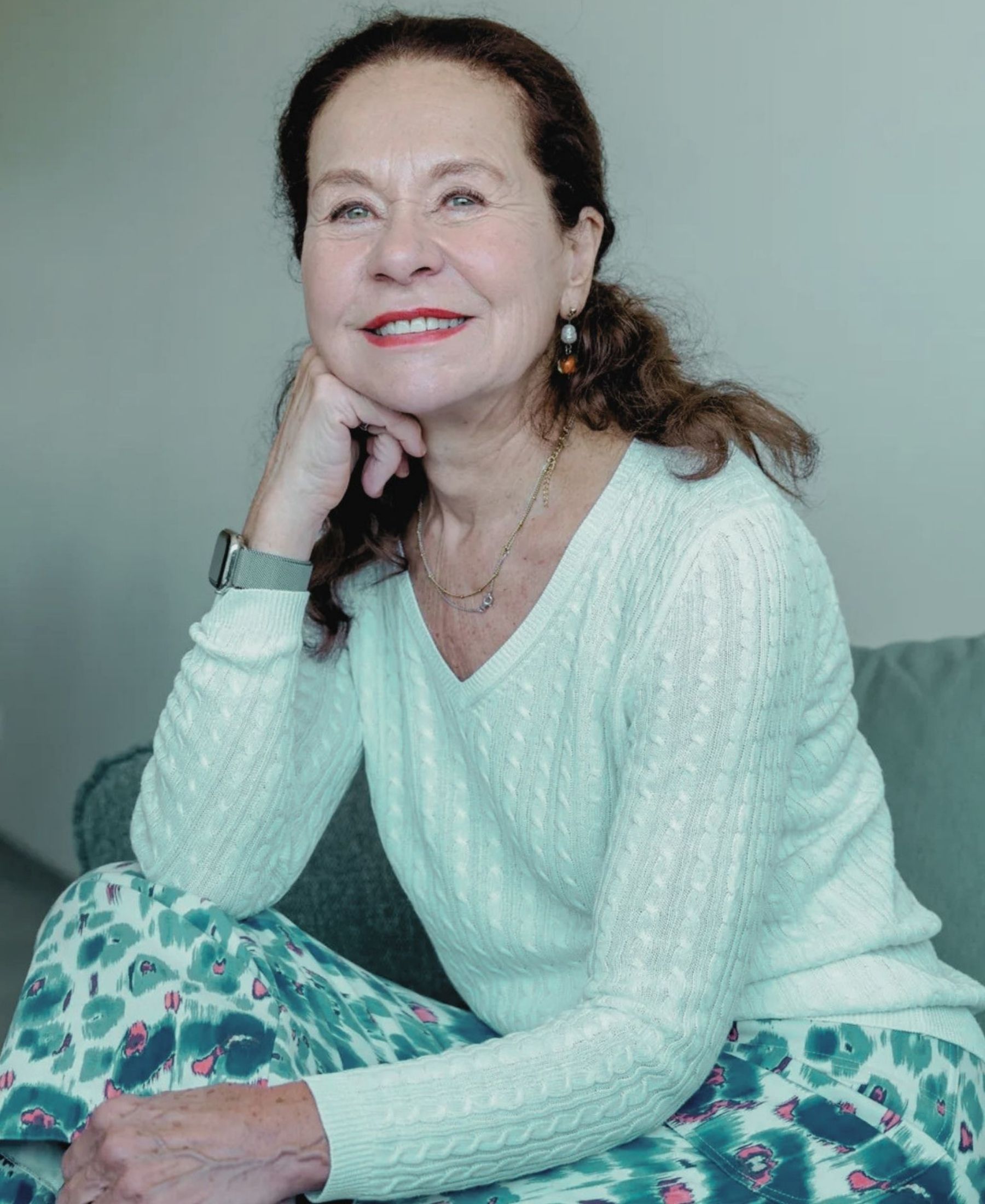Joëlle Ciocco seated and smiling, wearing a pale cable-knit sweater with patterned trousers.