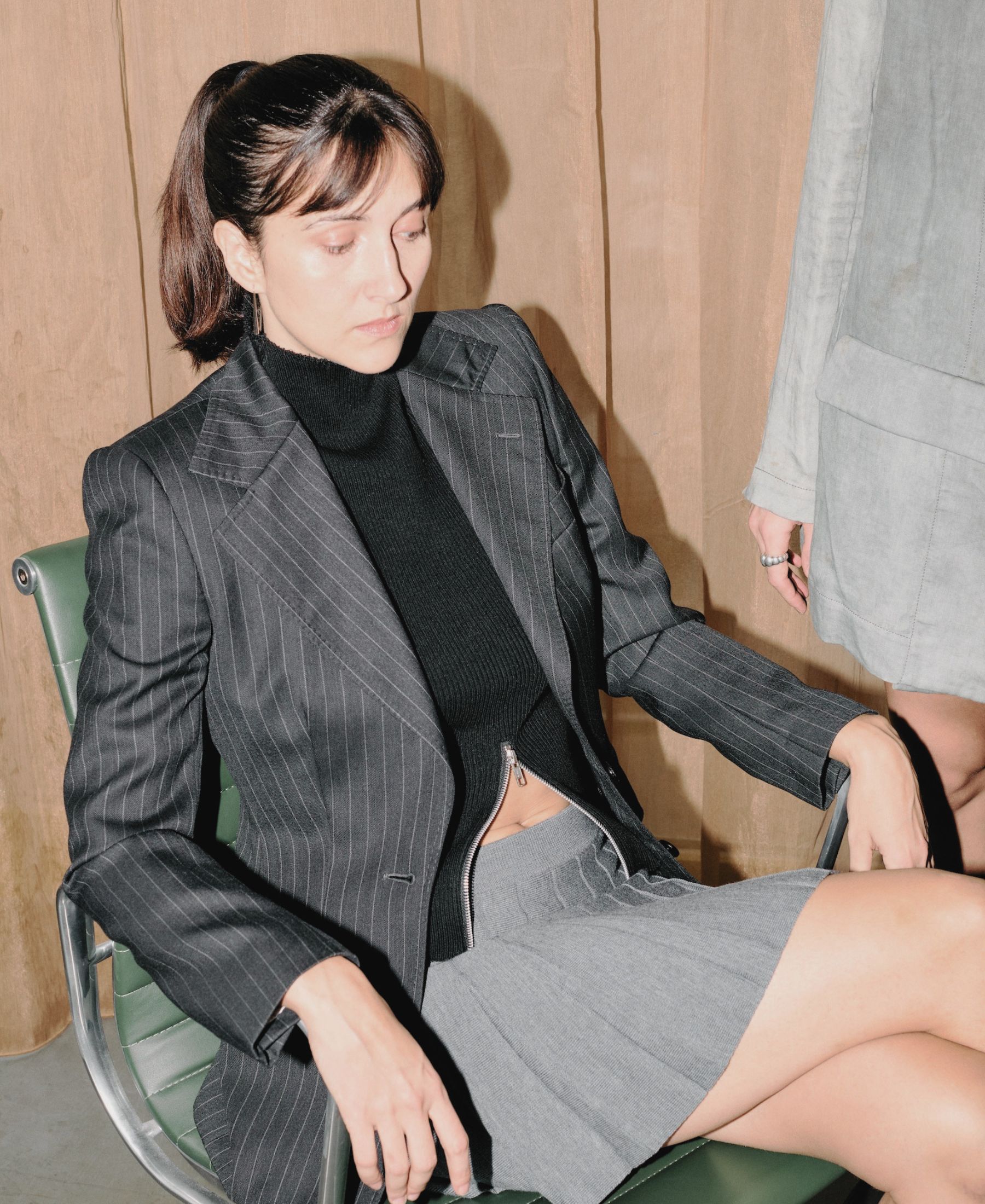 Kim Nüesch Sisters seated in a green chair, wearing a pinstripe blazer over a black knit and grey skirt.