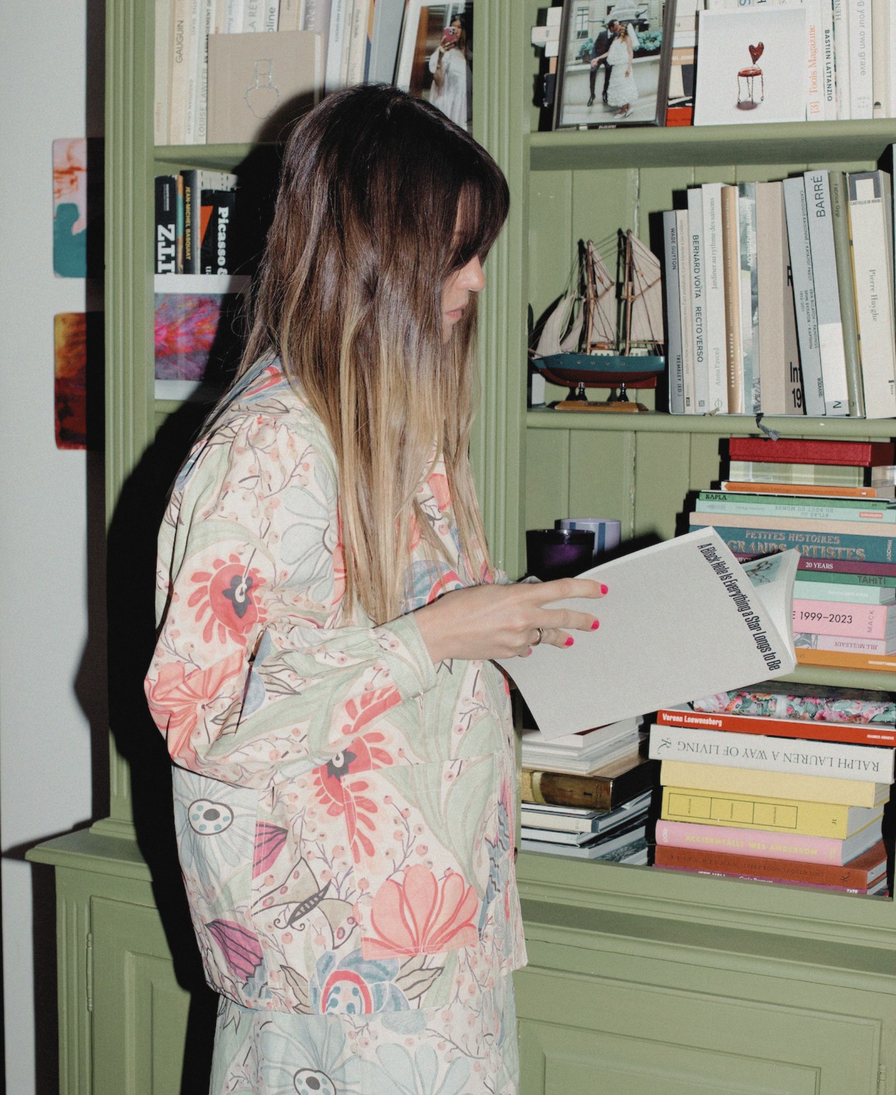 Anouck Mutsaerts blättert neben einem grünen Bücherregal mit Büchern, Fotografien und gesammelten Objekten in einem Kunstband.