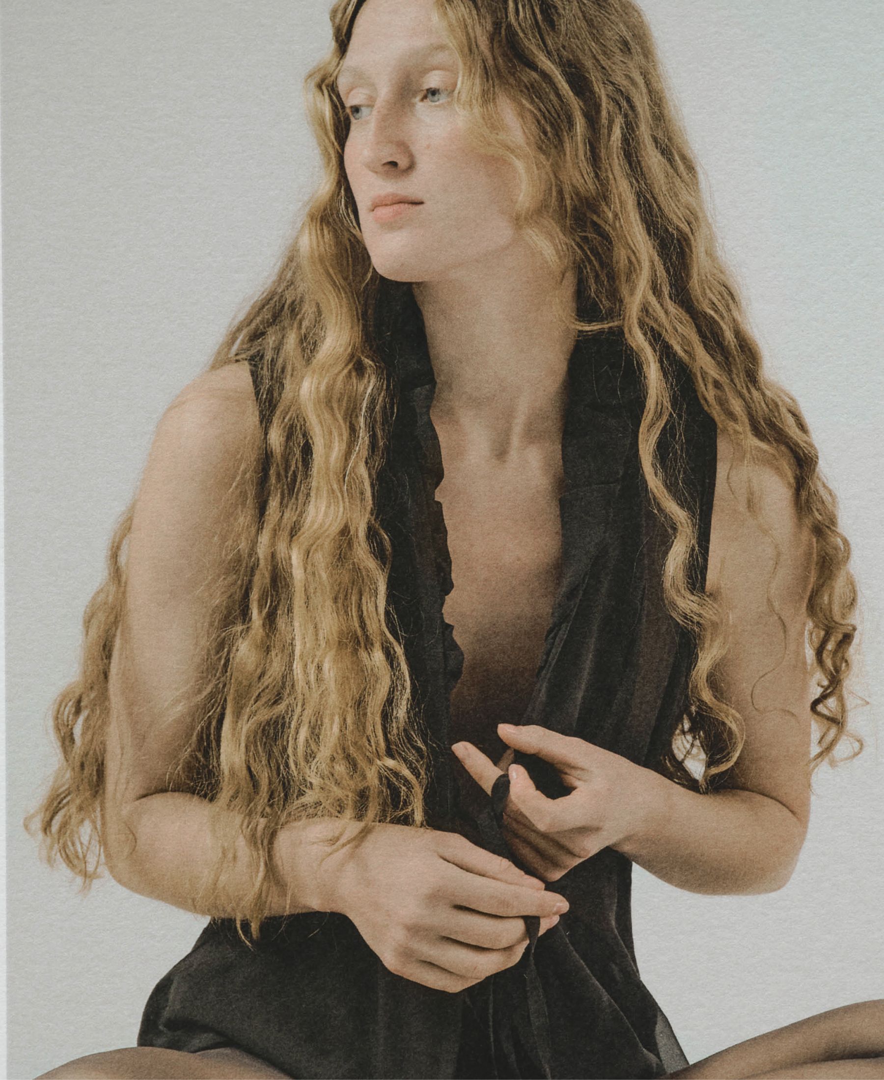 Sasha Konograeva with long wavy hair, seated in a black sleeveless look against a pale studio backdrop.