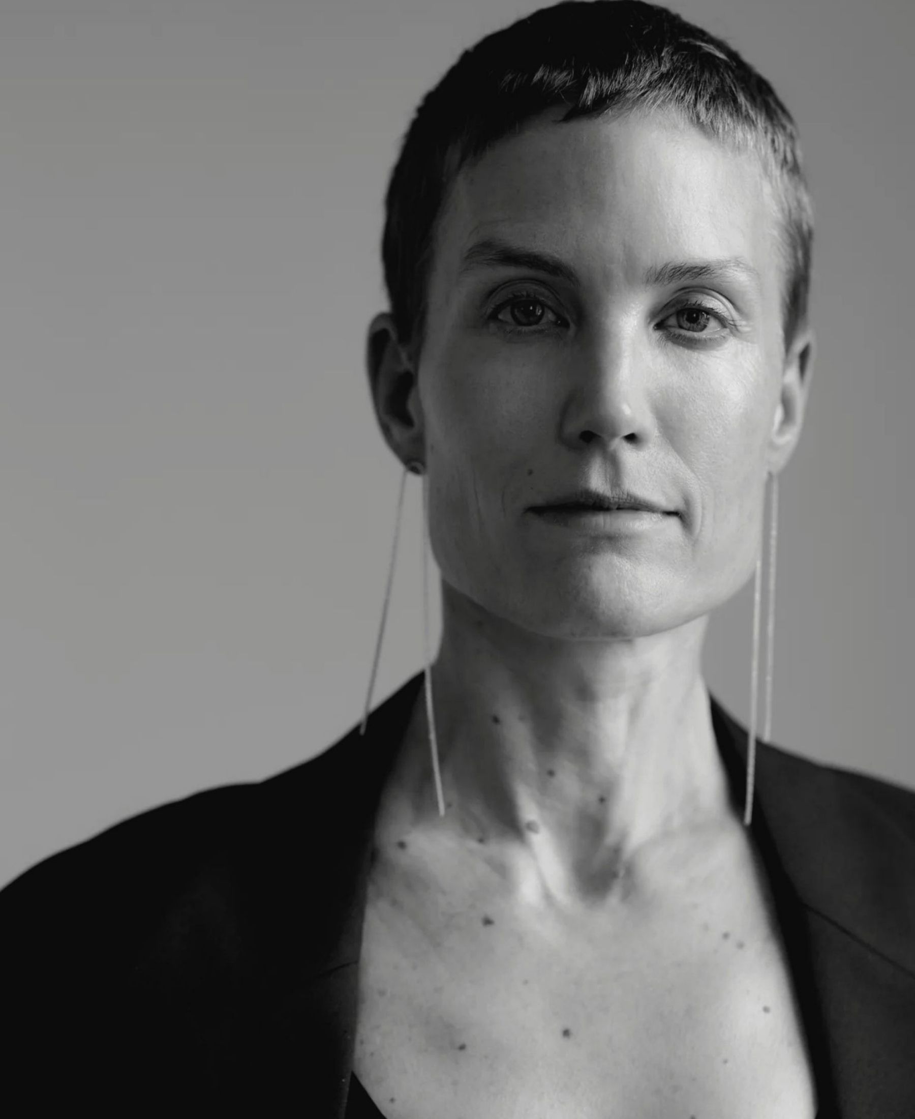 Black-and-white portrait of Saskia Diez with closely cropped hair, wearing a dark jacket and long fine earrings.