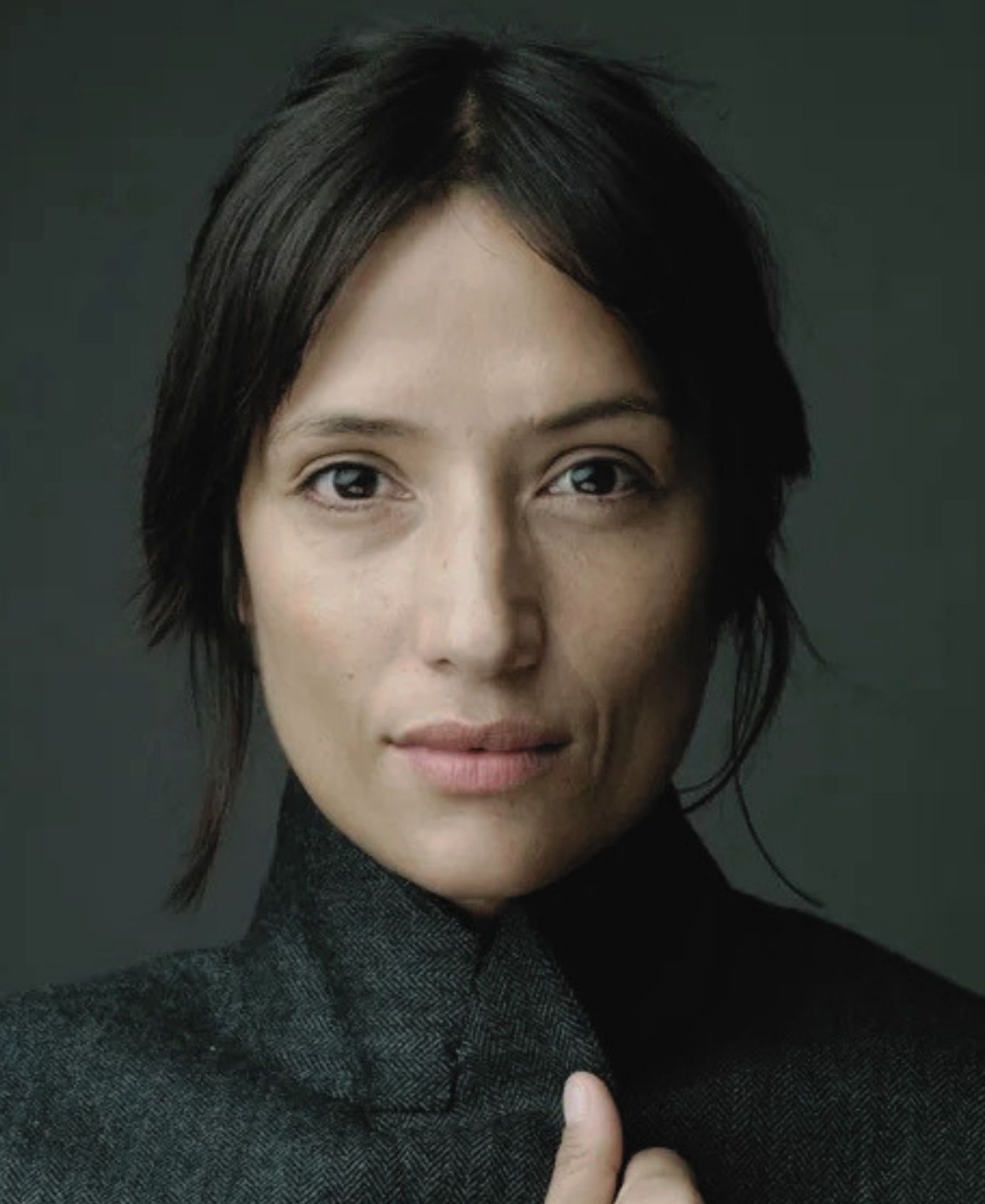 Close portrait of Melanie Winiger with dark hair loosely tied back, wearing a structured charcoal high-collared coat.