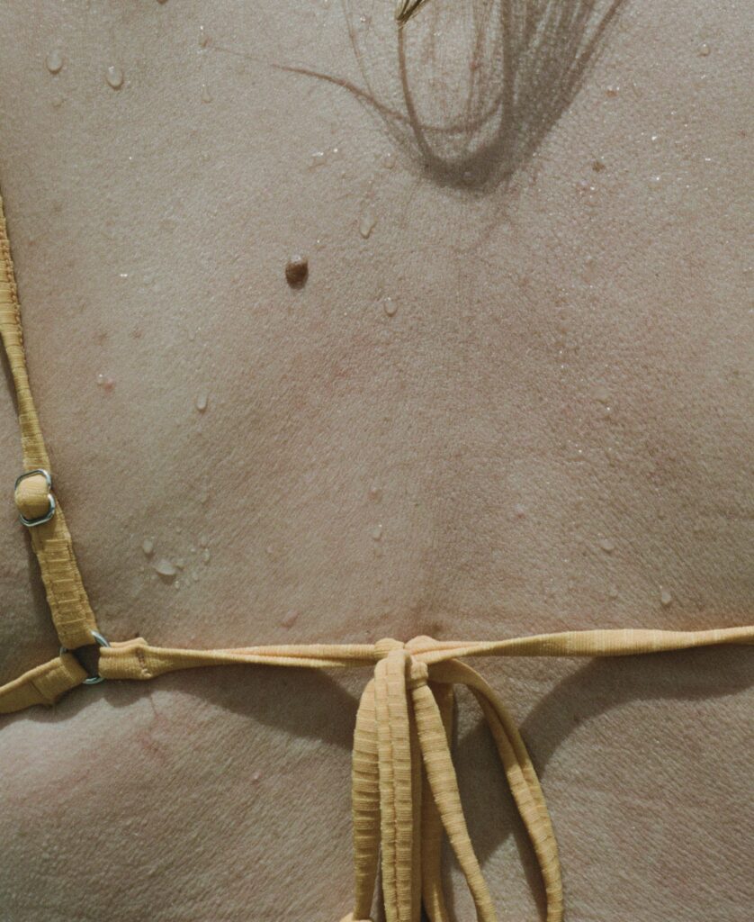 Close-up of sun-warmed skin dotted with water droplets, with the ties of a yellow bikini crossing the back.